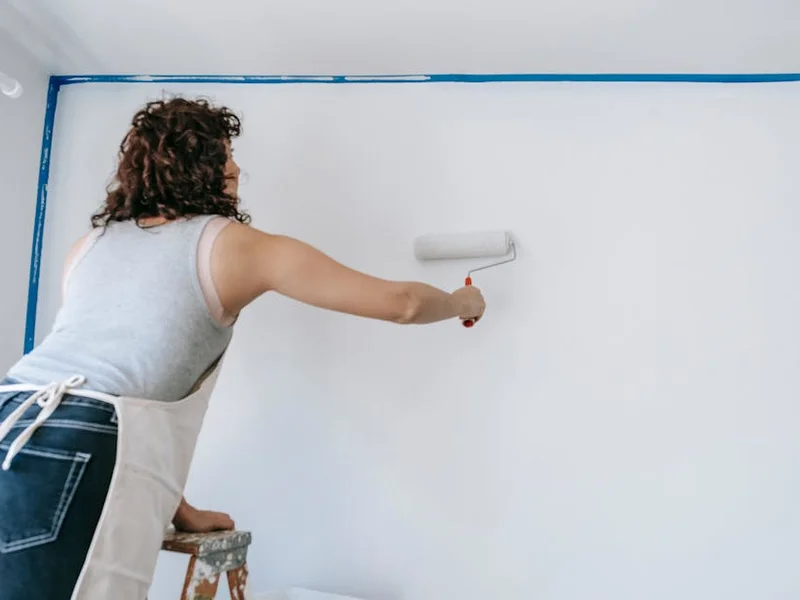 A woman stands on a ladder and paints a white wall during a home renovation project.