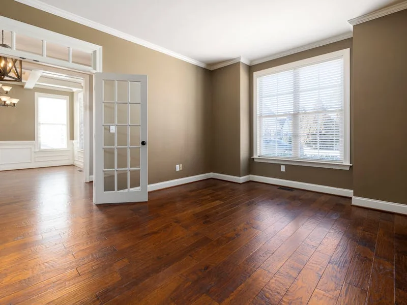 Bright and empty room with hardwood flooring, large windows, and elegant lighting.