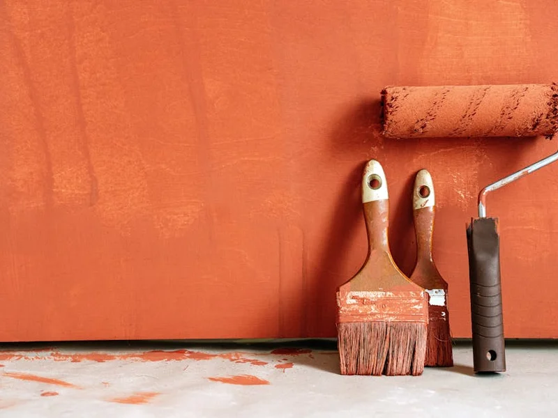 Close-up of paint rollers and brushes with orange paint against a painted wall.