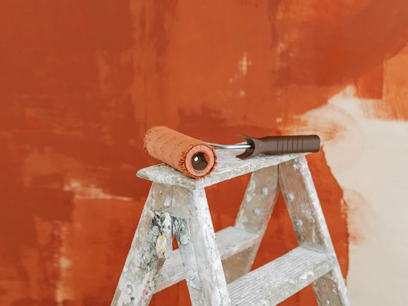 Paint roller on a ladder with a partially painted red wall in the background.