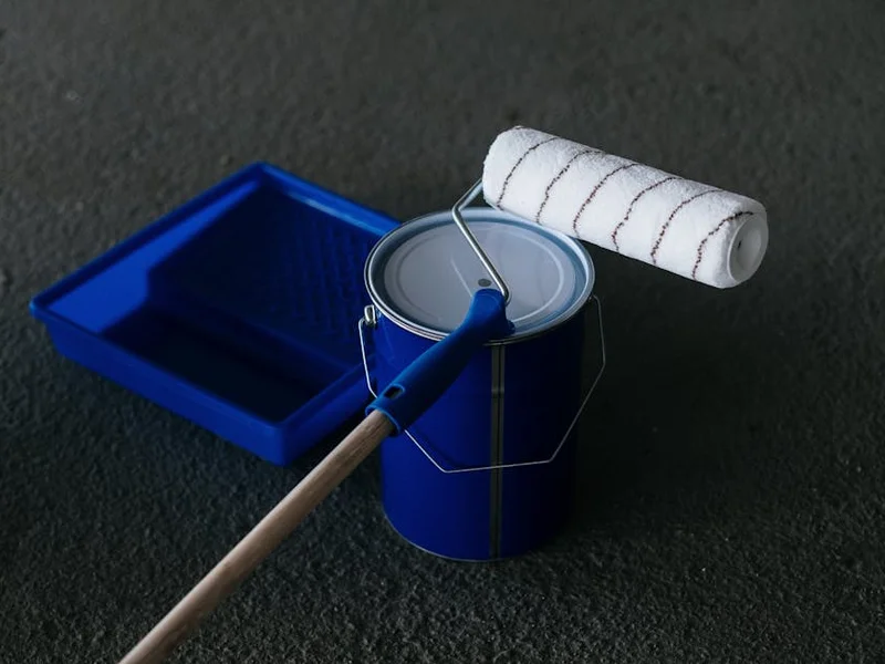Close-up of a paint can, roller, and tray on a textured floor, perfect for DIY projects.