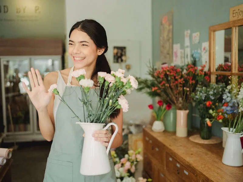 Friendly young florist waving in her shop, holding a bouquet of flowers.